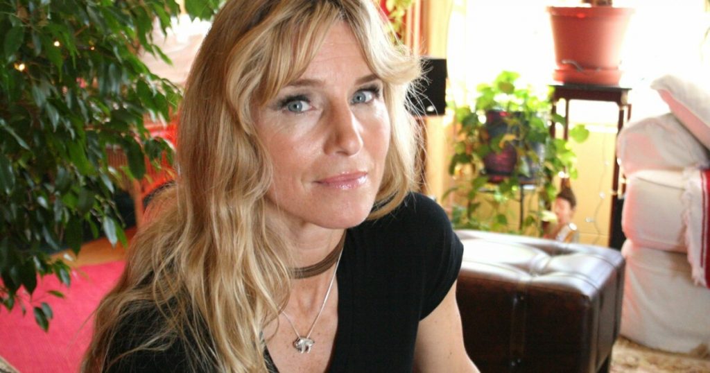 Portrait d’une femme blonde aux yeux clairs, assise dans un intérieur lumineux entouré de plantes vertes, regard direct vers l’objectif, portant un haut noir et un collier discret, ambiance chaleureuse et naturelle.