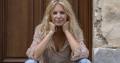 Portrait d’une femme blonde d’environ 60 ans, assise devant une porte en bois et un mur en pierre, les mains posées sous le menton, regard serein et léger sourire, vêtue d’une blouse claire et d’un jean, ambiance naturelle et lumineuse.