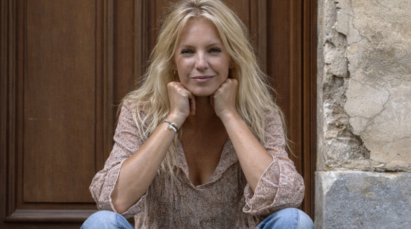 Portrait d’une femme blonde d’environ 60 ans, assise devant une porte en bois et un mur en pierre, les mains posées sous le menton, regard serein et léger sourire, vêtue d’une blouse claire et d’un jean, ambiance naturelle et lumineuse.
