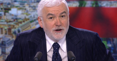 Présentateur de télévision aux cheveux blancs et barbe courte, vêtu d’un costume bleu marine à fines rayures et d’une cravate à pois, s’exprimant en direct derrière deux micros sur un plateau d’émission d’information, avec un fond flouté aux tons rouges et une vue urbaine en arrière-plan.