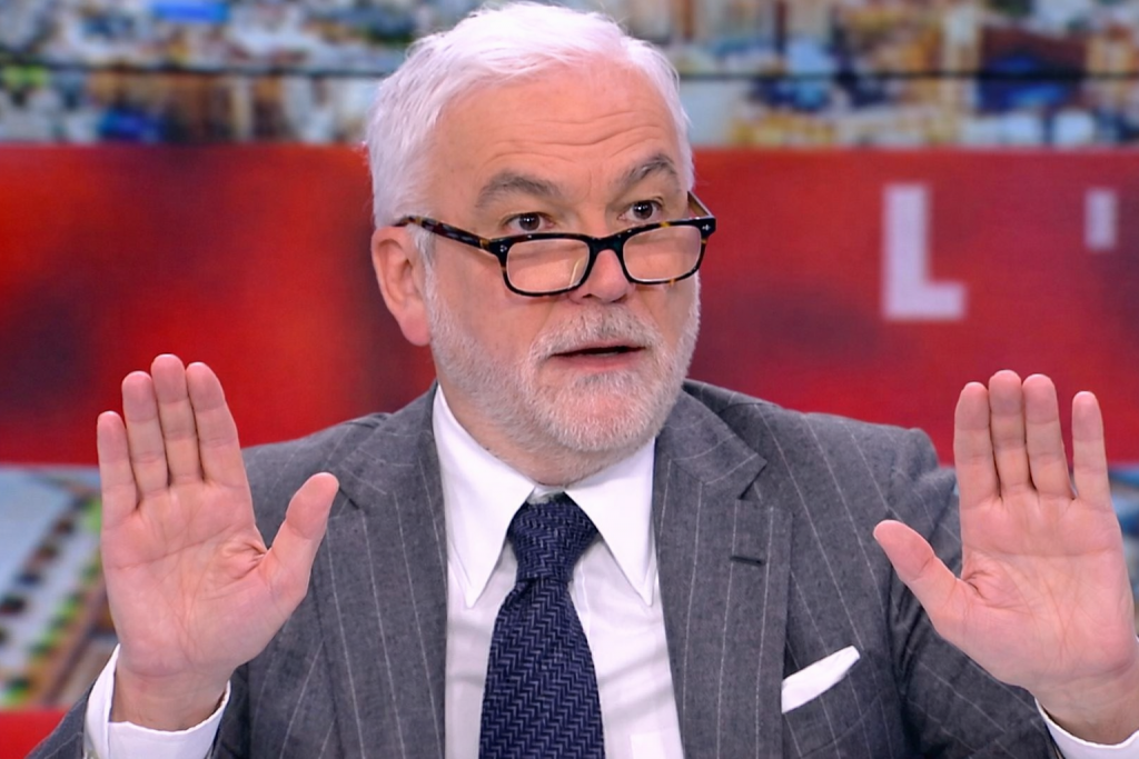 Animateur de télévision aux cheveux blancs portant des lunettes rectangulaires noires, vêtu d’un costume gris à fines rayures et d’une cravate bleu foncé, levant les deux mains paumes ouvertes en plein direct sur un plateau d’information, avec un fond rouge et une vue urbaine floutée en arrière-plan.