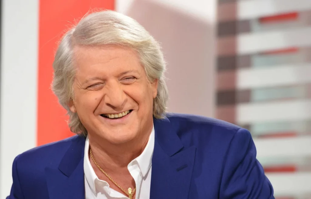 homme aux cheveux gris souriant en plateau de télévision, vêtu d’un costume bleu.