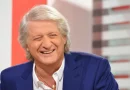 homme aux cheveux gris souriant en plateau de télévision, vêtu d’un costume bleu.