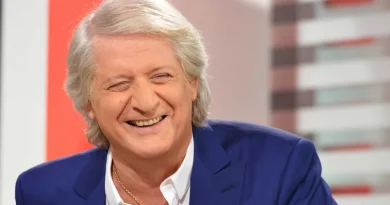 homme aux cheveux gris souriant en plateau de télévision, vêtu d’un costume bleu.