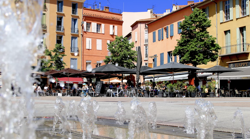Vue panoramique d’une ville du sud de la France baignée de soleil, avec des toits aux tuiles rouges, des façades claires, des palmiers et des montagnes en arrière-plan sous un ciel bleu dégagé.
