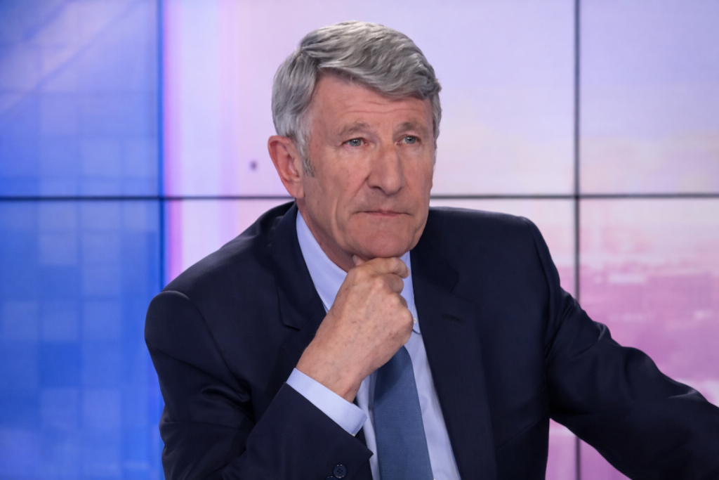 Un homme aux cheveux gris, assis sur un plateau de télévision, adopte une posture grave et réfléchie, la main posée sous le menton, dans une ambiance de débat télévisé tendu.