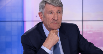 Un homme aux cheveux gris, assis sur un plateau de télévision, adopte une posture grave et réfléchie, la main posée sous le menton, dans une ambiance de débat télévisé tendu.