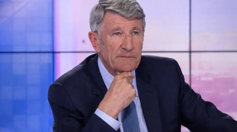 Un homme aux cheveux gris, assis sur un plateau de télévision, adopte une posture grave et réfléchie, la main posée sous le menton, dans une ambiance de débat télévisé tendu.