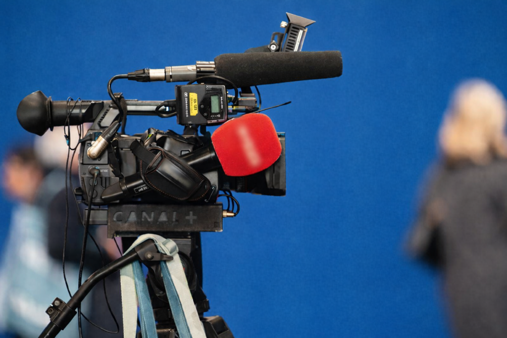 Caméra de télévision professionnelle installée sur un plateau, avec microphone recouvert de mousse, fond bleu et silhouettes floues en arrière-plan.