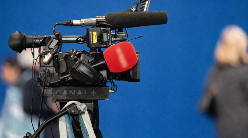 Caméra de télévision professionnelle installée sur un plateau, avec microphone recouvert de mousse, fond bleu et silhouettes floues en arrière-plan.