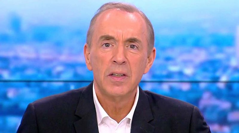 Présentateur masculin en costume sombre et chemise blanche, regard sérieux face caméra, en plateau de chaîne d’information avec arrière-plan flouté aux tons bleus représentant une ville.