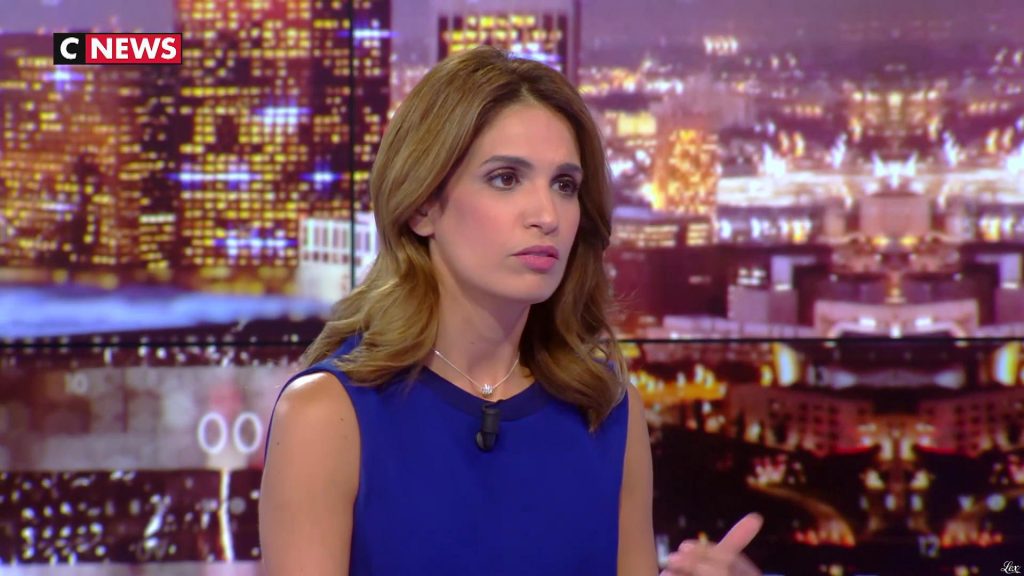 Présentatrice de télévision en plateau d’information, tenue bleue, regard concentré, décor urbain nocturne flou en arrière-plan