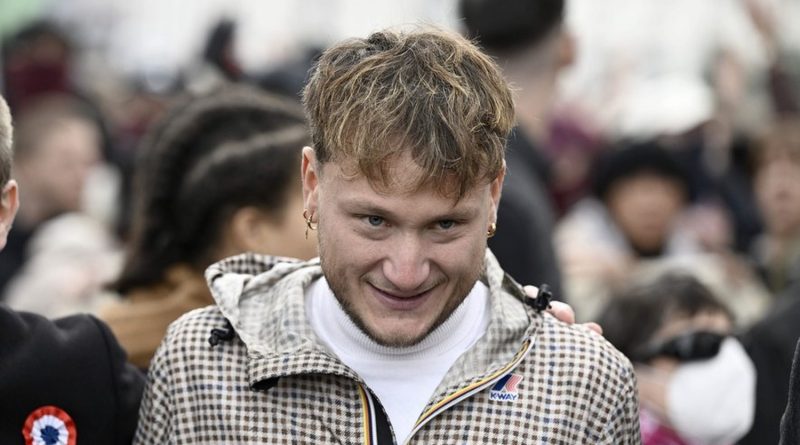 Jeune homme aux cheveux courts et clairs, portant une veste à motifs beige et marron et un col roulé blanc, esquissant un léger sourire au milieu d’une foule rassemblée en extérieur.