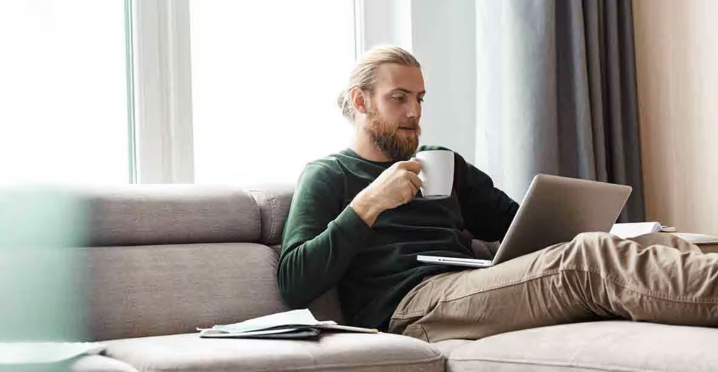 Homme adulte blond avec barbe courte, assis sur un canapé beige dans un salon lumineux, tenant une tasse blanche tout en utilisant un ordinateur portable posé sur ses jambes. Il porte un pull vert foncé et un pantalon beige. À côté de lui, un carnet ouvert est visible sur le canapé. Une grande fenêtre laisse entrer la lumière naturelle, créant une atmosphère calme et chaleureuse. L’image illustre le télétravail, la gestion des finances personnelles ou la lecture d’informations en ligne dans un environnement domestique moderne et apaisant.