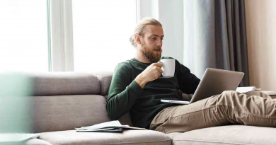Homme adulte blond avec barbe courte, assis sur un canapé beige dans un salon lumineux, tenant une tasse blanche tout en utilisant un ordinateur portable posé sur ses jambes. Il porte un pull vert foncé et un pantalon beige. À côté de lui, un carnet ouvert est visible sur le canapé. Une grande fenêtre laisse entrer la lumière naturelle, créant une atmosphère calme et chaleureuse. L’image illustre le télétravail, la gestion des finances personnelles ou la lecture d’informations en ligne dans un environnement domestique moderne et apaisant.