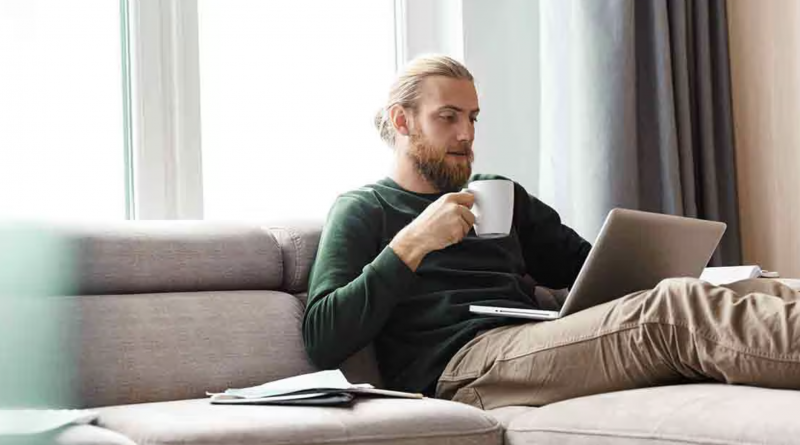Homme adulte blond avec barbe courte, assis sur un canapé beige dans un salon lumineux, tenant une tasse blanche tout en utilisant un ordinateur portable posé sur ses jambes. Il porte un pull vert foncé et un pantalon beige. À côté de lui, un carnet ouvert est visible sur le canapé. Une grande fenêtre laisse entrer la lumière naturelle, créant une atmosphère calme et chaleureuse. L’image illustre le télétravail, la gestion des finances personnelles ou la lecture d’informations en ligne dans un environnement domestique moderne et apaisant.