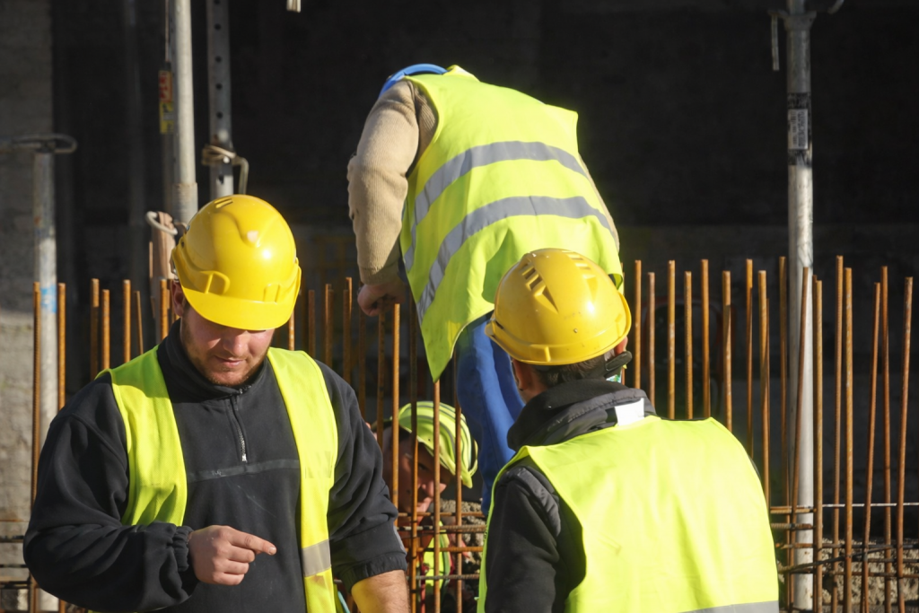 Ouvriers sur un chantier de construction, équipés de casques et de gilets de sécurité, travaillant autour d’armatures métalliques, illustrant une situation de travail et d’enquête sociale.