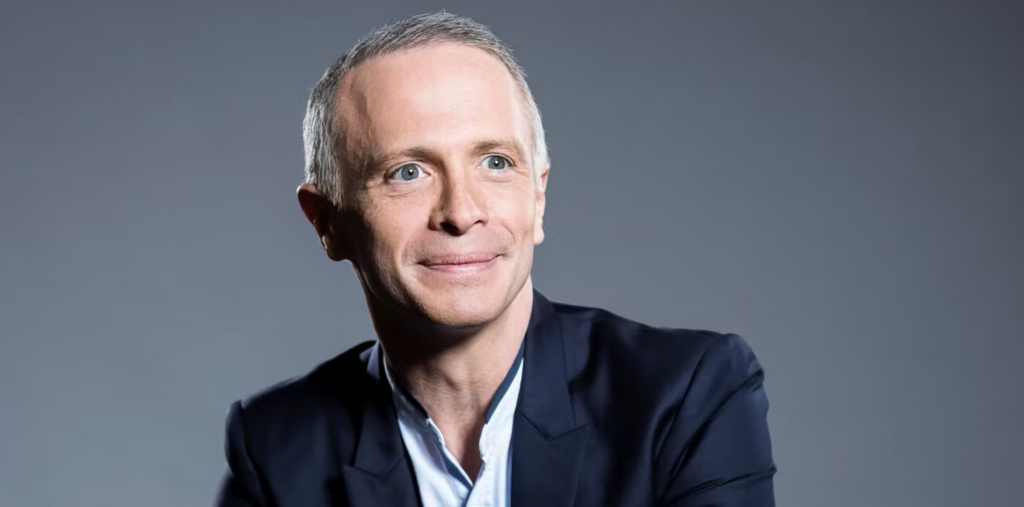 Animateur de télévision assis derrière un pupitre sur un plateau de jeu, veste sombre et chemise claire, sourire face caméra, décor lumineux composé de bandes colorées bleues et jaunes, ambiance studio moderne typique d’une émission de divertissement en France.