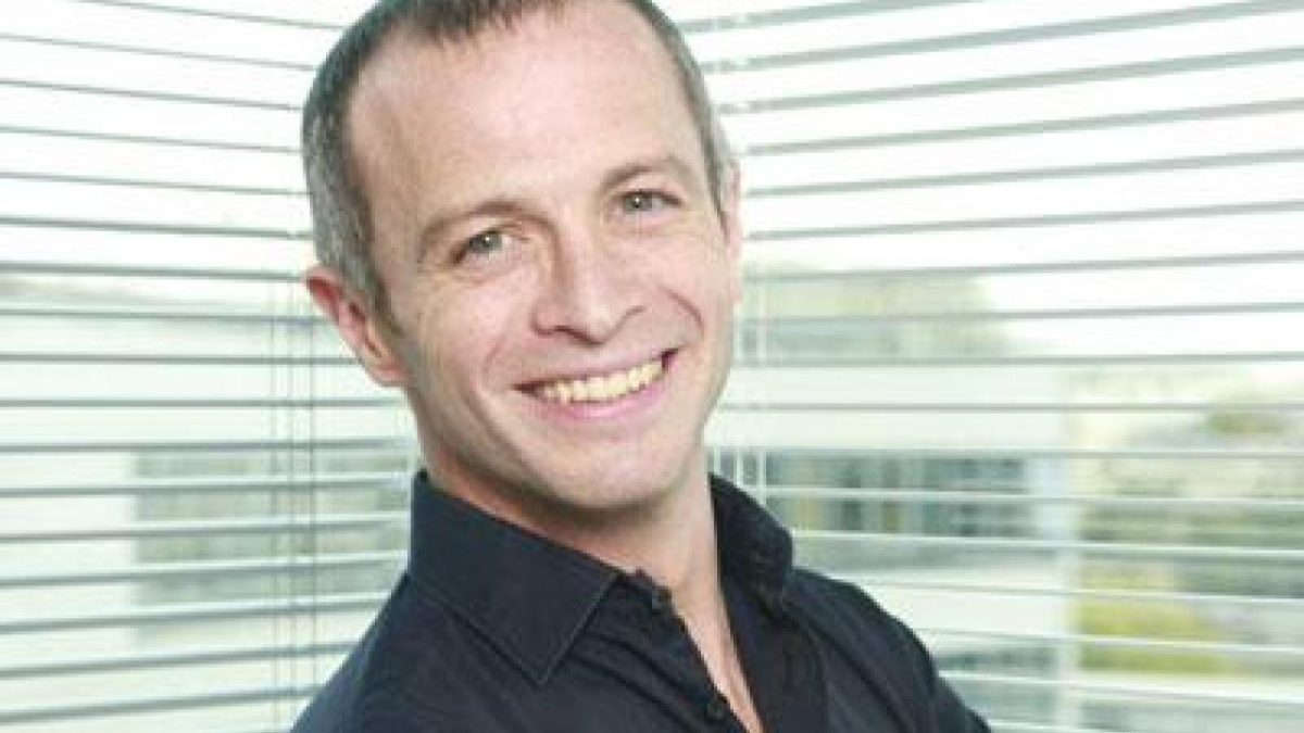 Portrait en plan rapproché d’un animateur de télévision souriant, cheveux courts grisonnants, chemise sombre, photographié devant une fenêtre avec stores horizontaux laissant passer la lumière naturelle, ambiance bureau ou studio lumineux.