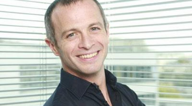 Portrait en plan rapproché d’un animateur de télévision souriant, cheveux courts grisonnants, chemise sombre, photographié devant une fenêtre avec stores horizontaux laissant passer la lumière naturelle, ambiance bureau ou studio lumineux.