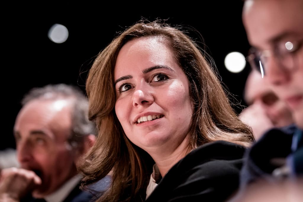 Une femme assise lors d’un événement public, regard tourné vers la scène, expression attentive, entourée d’autres personnes légèrement floutées.