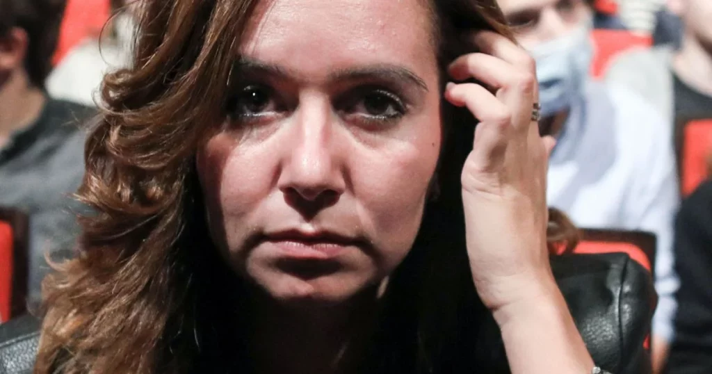 Gros plan sur une femme assise dans une salle, le visage fermé et concentré, la main près des cheveux, avec un public flou en arrière-plan.