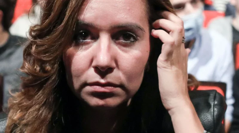 Gros plan sur une femme assise dans une salle, le visage fermé et concentré, la main près des cheveux, avec un public flou en arrière-plan.