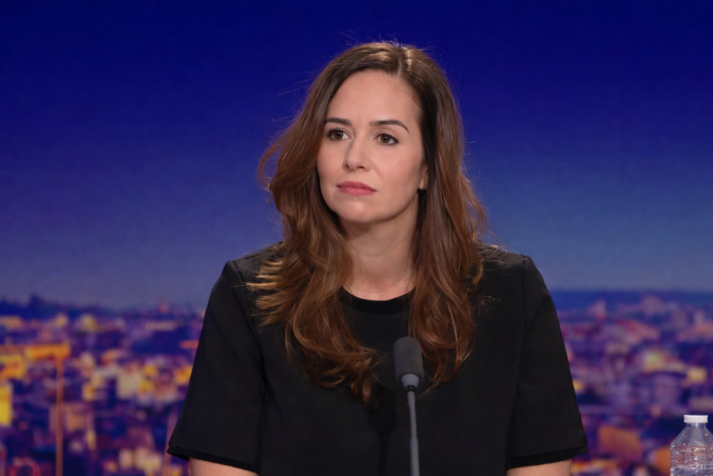 Une femme politique s’exprime face caméra lors d’une interview en plateau, avec un micro posé devant elle et un décor urbain flouté en arrière-plan.