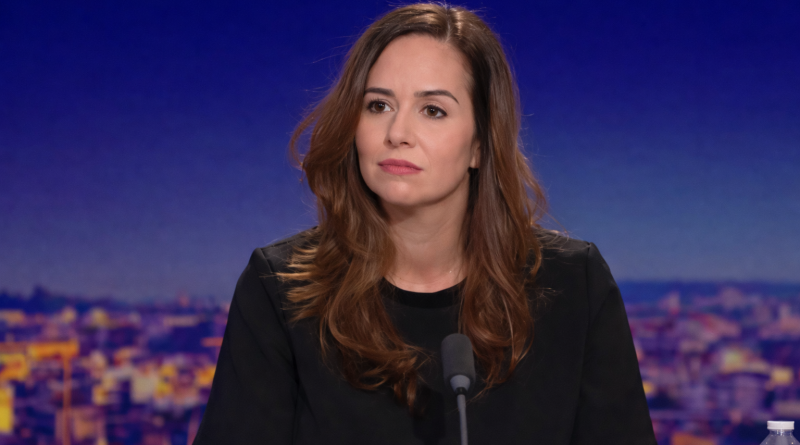 Une femme politique s’exprime face caméra lors d’une interview en plateau, avec un micro posé devant elle et un décor urbain flouté en arrière-plan.