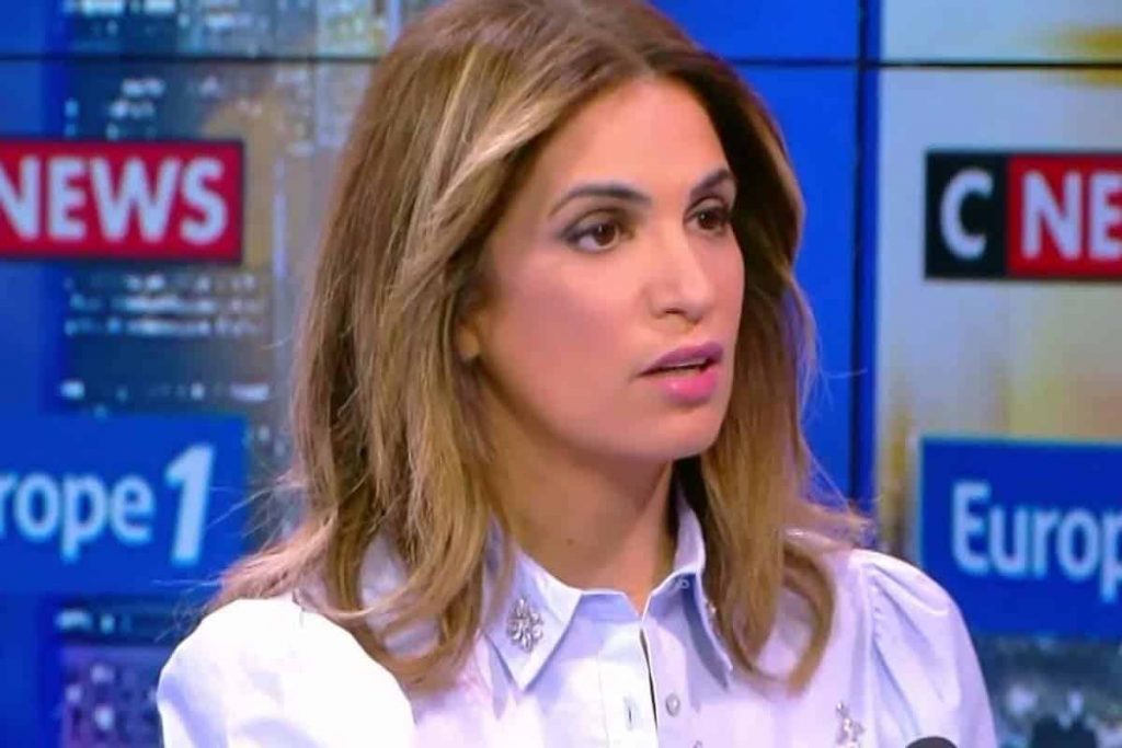 Journaliste brune aux cheveux mi-longs intervenant en plateau télévisé, vêtue d’une chemise claire, devant un décor d’émission d’information avec écrans et logos de médias en arrière-plan.