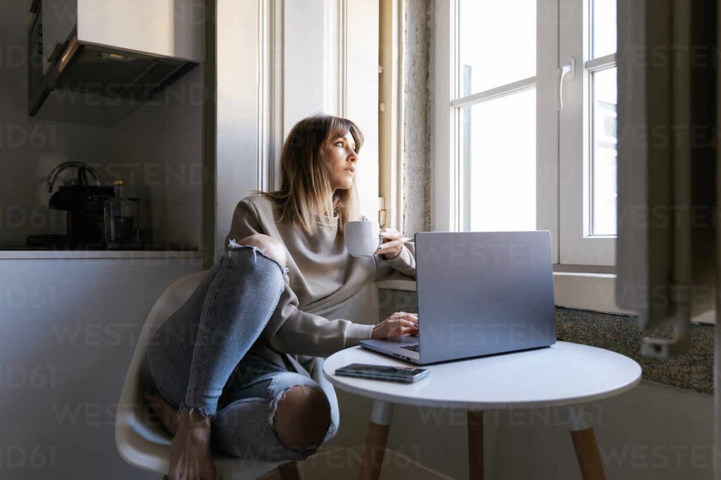 Femme adulte assise sur une chaise moderne près d’une grande fenêtre, travaillant sur un ordinateur portable posé sur une petite table ronde blanche. Elle tient une tasse blanche à la main et regarde vers l’extérieur, plongée dans ses pensées. Elle porte un pull beige ample et un jean bleu légèrement déchiré au genou. Un smartphone est posé sur la table à côté de l’ordinateur. La lumière naturelle entre abondamment par la fenêtre, créant une atmosphère douce, calme et introspective. L’image évoque le télétravail, la gestion des finances personnelles, la réflexion professionnelle ou la lecture d’actualités économiques dans un intérieur cosy et lumineux.