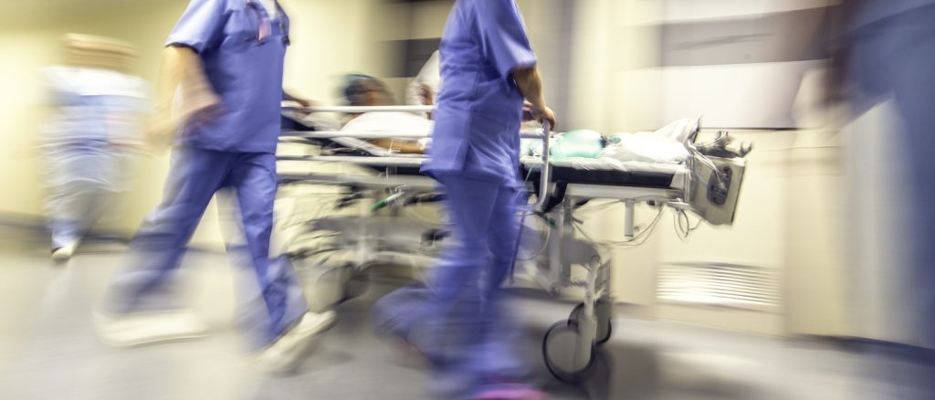 Personnel soignant transportant un patient sur un brancard dans un couloir d’hôpital, mouvement flou suggérant l’urgence, ambiance de tension et de prise en charge médicale rapide.