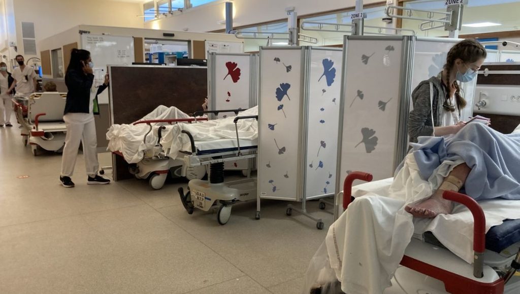 Couloir d’un service d’urgences hospitalières avec plusieurs brancards alignés, patients allongés derrière des paravents médicaux, personnel soignant en mouvement, atmosphère de saturation et d’attente prolongée.