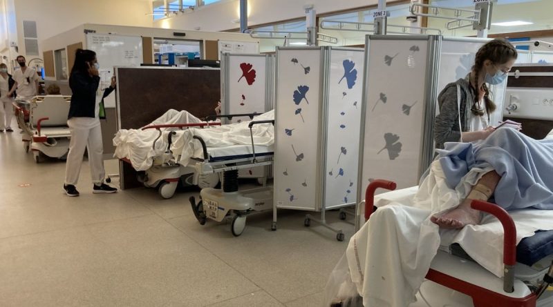 Couloir d’un service d’urgences hospitalières avec plusieurs brancards alignés, patients allongés derrière des paravents médicaux, personnel soignant en mouvement, atmosphère de saturation et d’attente prolongée.