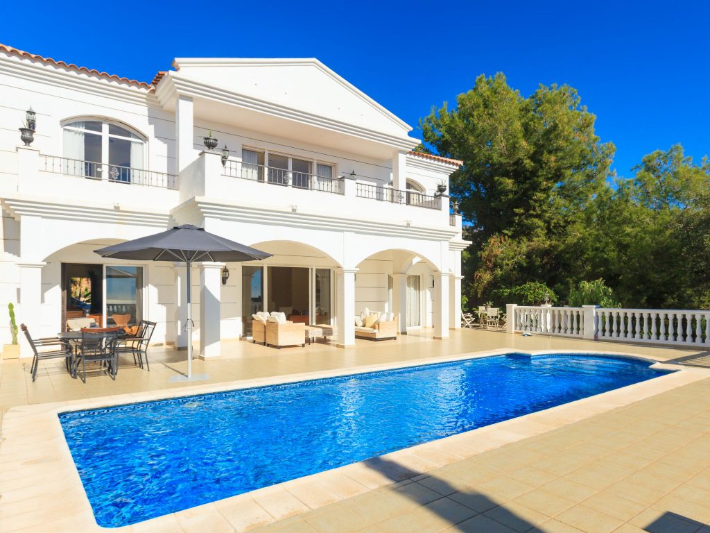 Grande villa blanche de style méditerranéen avec deux étages, terrasse spacieuse, piscine rectangulaire d’un bleu intense au premier plan, mobilier de jardin et parasol, entourée d’arbres verdoyants sous un ciel parfaitement dégagé.
