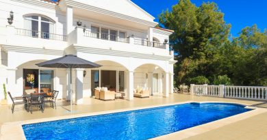 Grande villa blanche de style méditerranéen avec deux étages, terrasse spacieuse, piscine rectangulaire d’un bleu intense au premier plan, mobilier de jardin et parasol, entourée d’arbres verdoyants sous un ciel parfaitement dégagé.