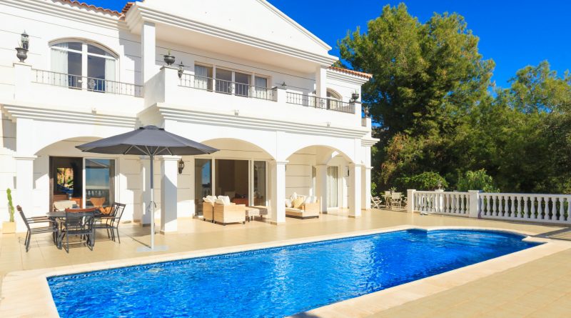 Grande villa blanche de style méditerranéen avec deux étages, terrasse spacieuse, piscine rectangulaire d’un bleu intense au premier plan, mobilier de jardin et parasol, entourée d’arbres verdoyants sous un ciel parfaitement dégagé.