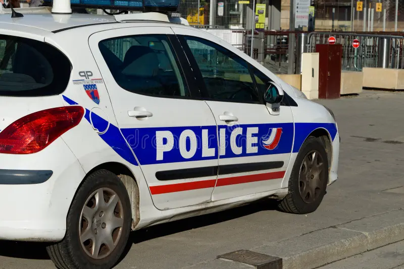 Voiture de police blanche avec marquage bleu et rouge stationnée sur la voie publique en milieu urbain, gyrophares visibles sur le toit, bâtiments et barrières en arrière-plan.