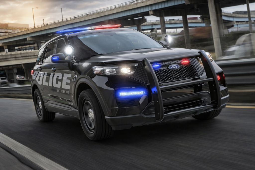 Véhicule de police noir équipé de gyrophares rouges et bleus allumés circulant rapidement sur une autoroute urbaine, avec échangeurs routiers en béton et circulation floutée en arrière-plan.
