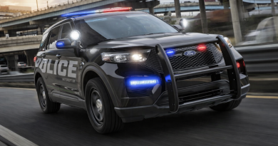 Véhicule de police noir équipé de gyrophares rouges et bleus allumés circulant rapidement sur une autoroute urbaine, avec échangeurs routiers en béton et circulation floutée en arrière-plan.