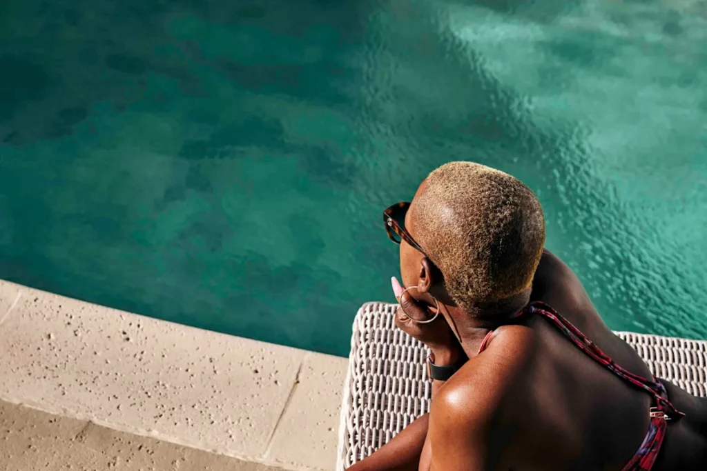 Une personne allongée sur un transat au bord d’une piscine à l’eau turquoise, profitant du soleil dans une atmosphère de détente et de bien-être.