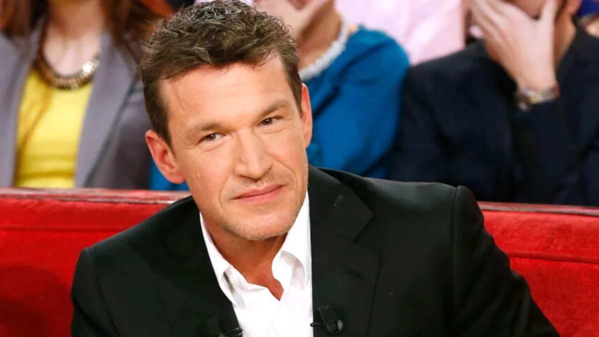Animateur de télévision français assis sur un canapé rouge lors d’une émission télévisée, vêtu d’une veste noire et d’une chemise blanche, regard tourné vers la caméra avec une expression sérieuse, tandis qu’un public flou apparaît en arrière-plan sur un plateau de télévision lumineux.