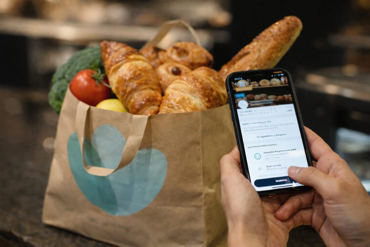 Personne tenant un smartphone affichant une application de récupération de nourriture invendue, devant un sac rempli de produits alimentaires comme du pain et des viennoiseries dans une boulangerie en fin de journée.