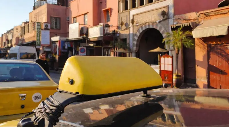 Taxi jaune stationné dans une rue animée d’une grande ville nord-africaine avec bâtiments traditionnels, boutiques et passants dans un quartier touristique en plein jour.
