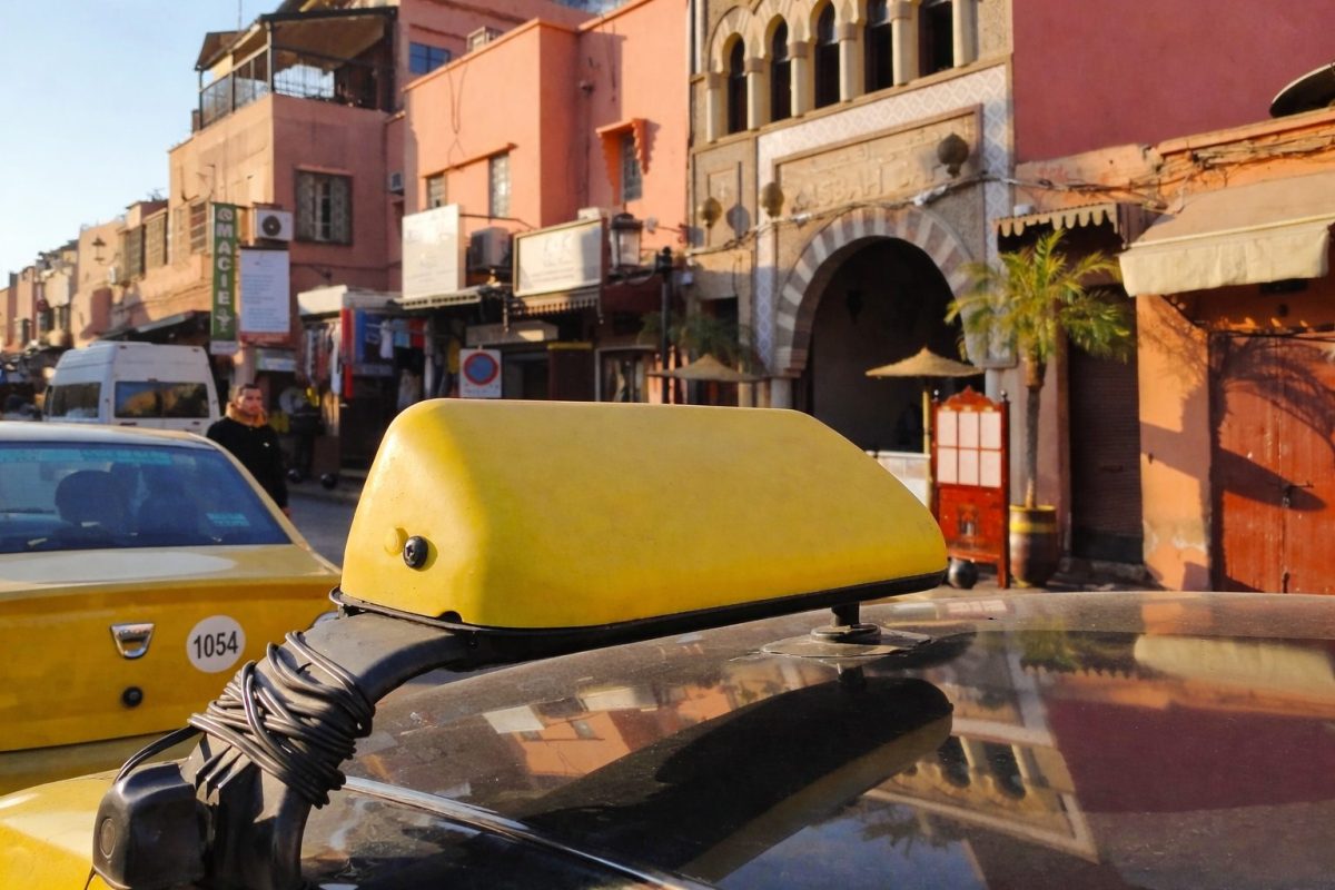 Taxi jaune stationné dans une rue animée d’une grande ville nord-africaine avec bâtiments traditionnels, boutiques et passants dans un quartier touristique en plein jour.