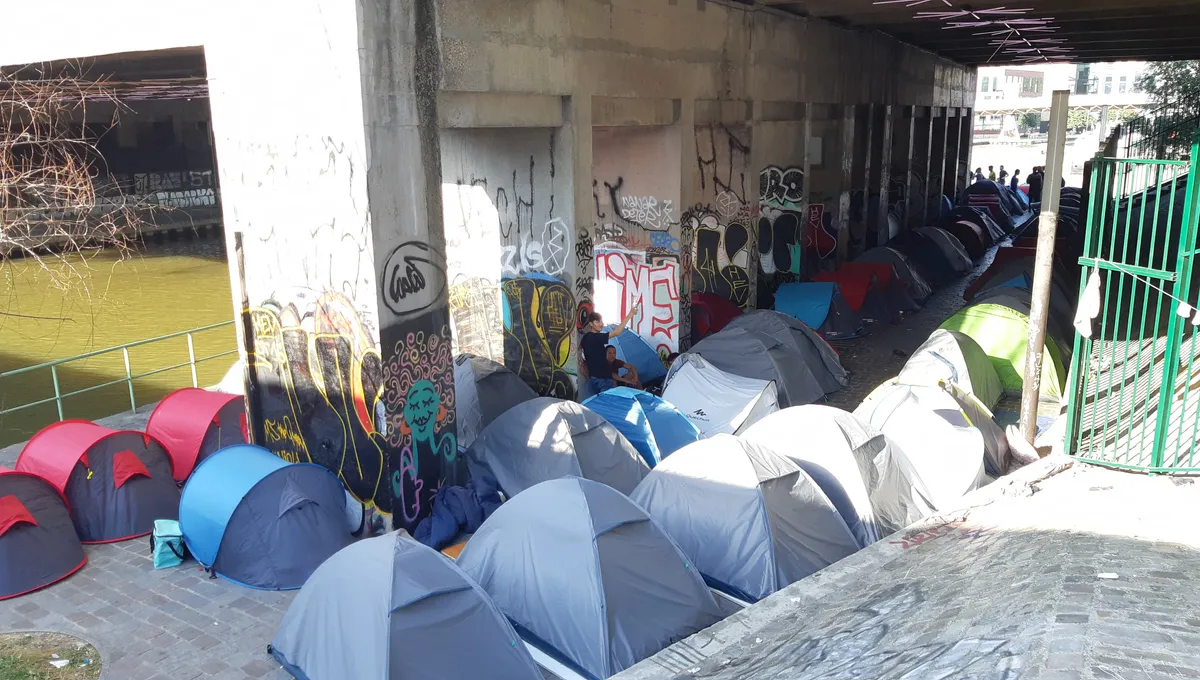 campement de tentes installé dans une rue étroite d’une ville avec plusieurs personnes vivant sur place illustrant la précarité et le débat sur l’accueil des migrants