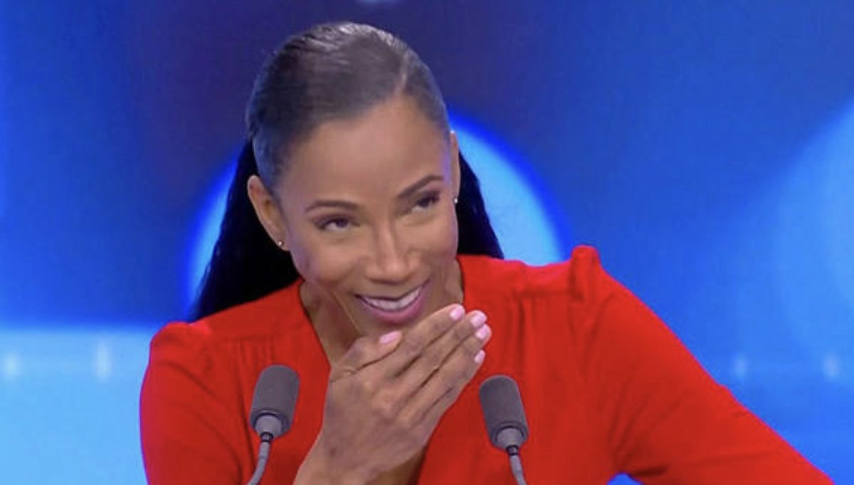 Présentatrice souriante en plateau télé, main devant la bouche dans un moment spontané en direct, ambiance studio avec éclairage professionnel