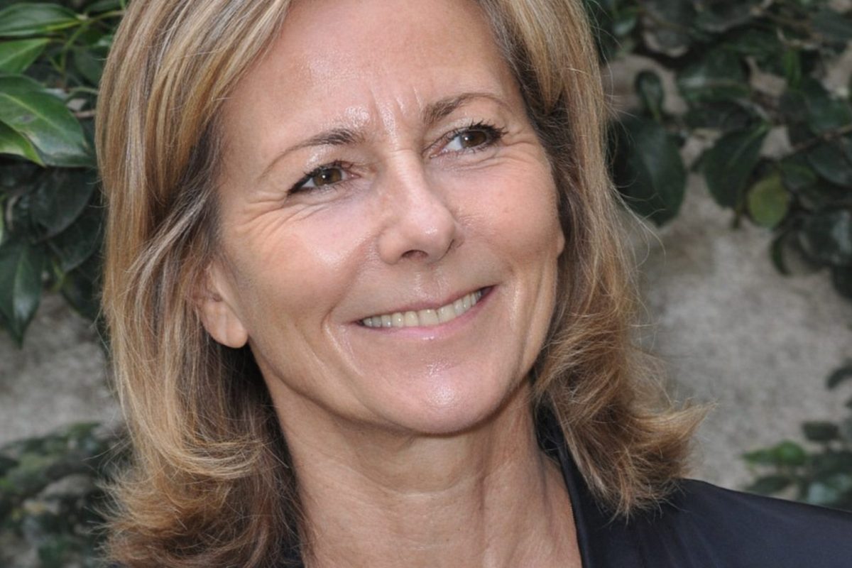 portrait en gros plan d’une femme blonde souriante lors d’un événement public, visage éclairé par une lumière naturelle avec un fond végétal vert légèrement flou