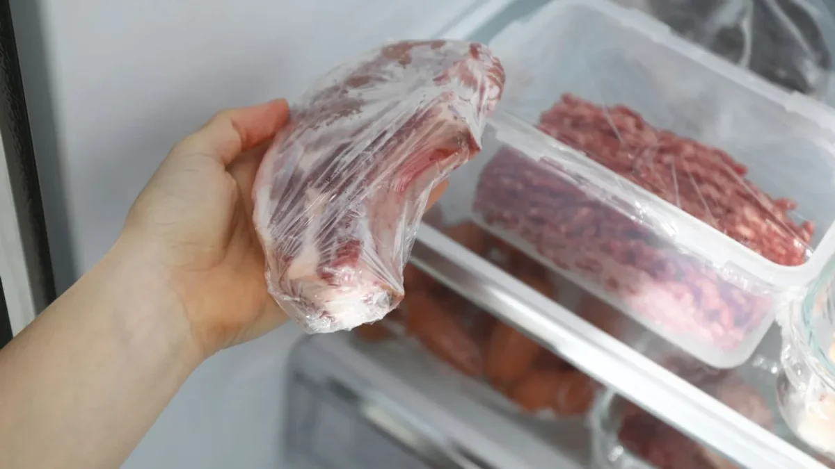 main plaçant un morceau de viande crue emballé dans un congélateur domestique avec d’autres aliments stockés