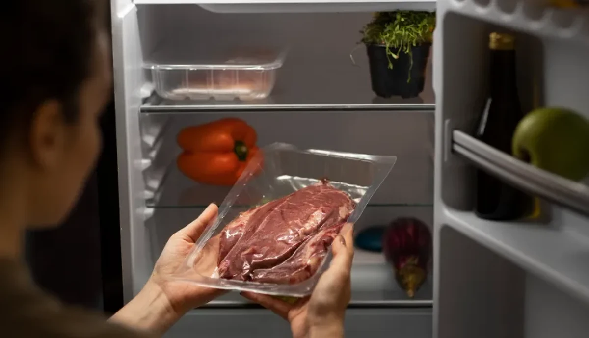 personne tenant un morceau de viande crue emballé devant un réfrigérateur ouvert avec des aliments à l’intérieur
